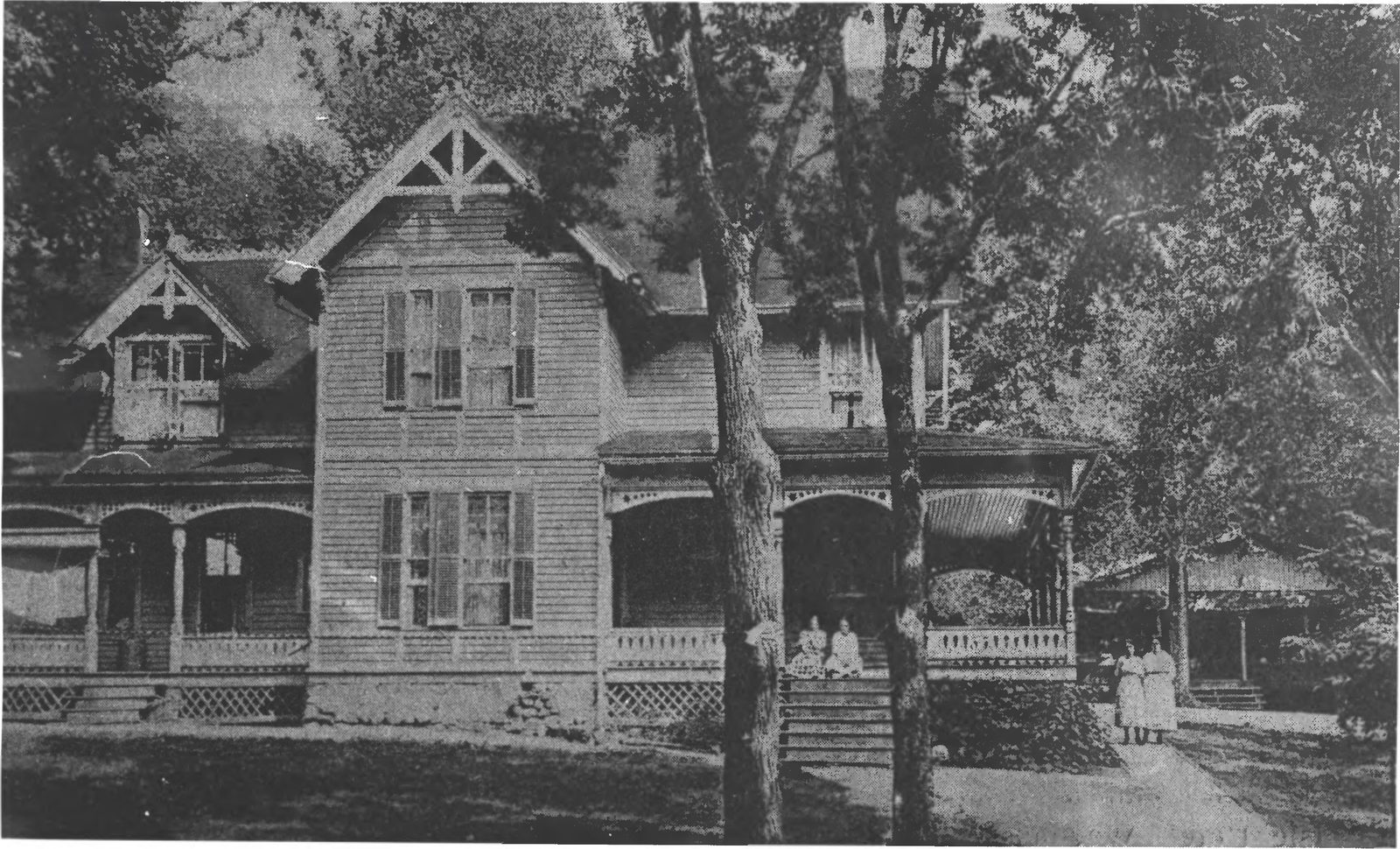 Historic photo of a large Victorian frame hotel on Coney Island in Lake Waconia, with guests sitting on the front steps and two women in white on the path