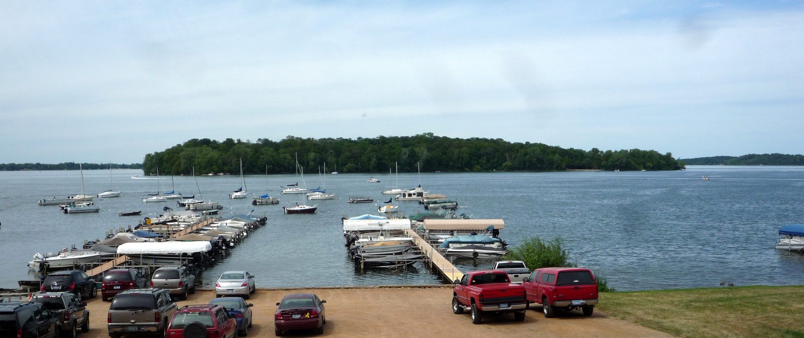 Coney Island seen across Lake Waconia beyond a Waconia shoreline marina with docked boats and a parking lot