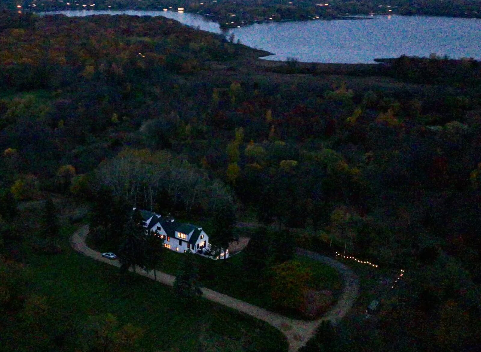 The Ches Mar Homestead house lit up at dusk on a fall evening, the curved gravel drive lined with lights, and Lake Minnewashta beyond