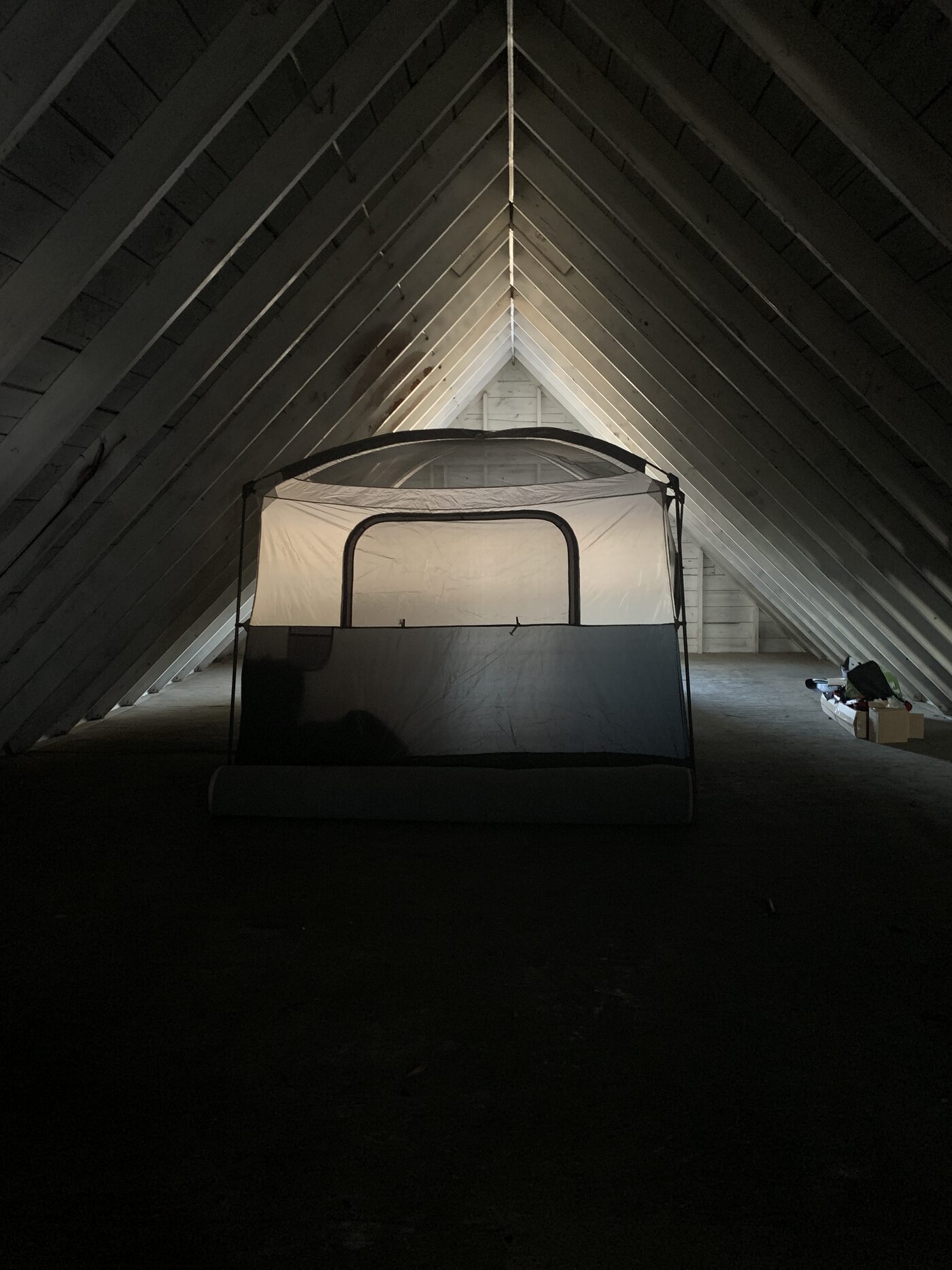 A camping tent set up over a bed inside the unfinished, windowless attic with its peaked wooden rafters